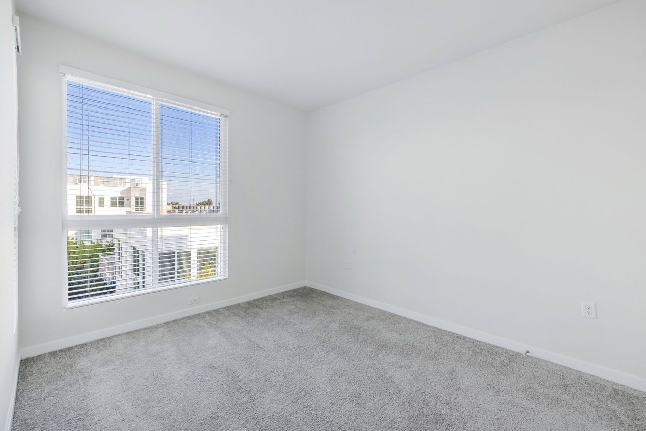 Empty bedroom with white walls, a large window with blinds, and carpet.