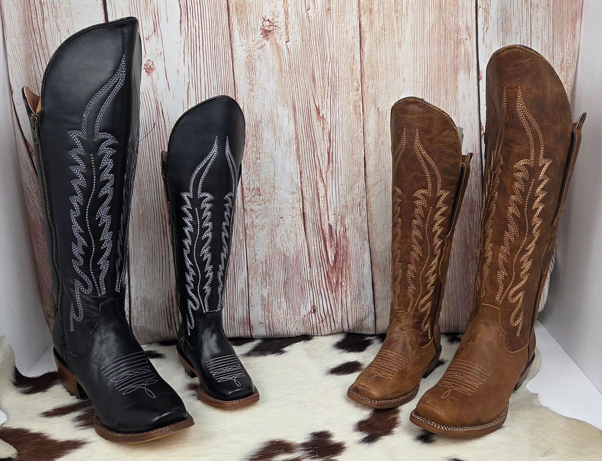 Pair of black and brown embroidered cowboy boots displayed on a patterned rug against a rustic wood backdrop