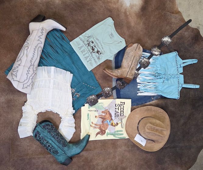 Assorted western-style clothing and accessories laid out on brown fabric: boots, hats, dresses, scarf, and book.