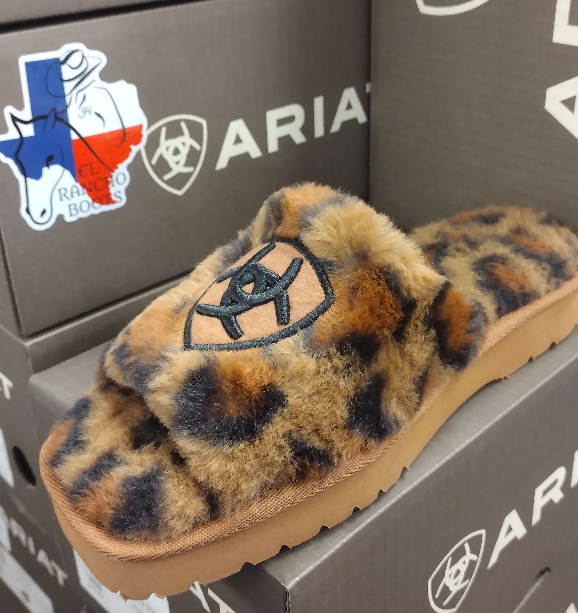 Leopard-print fuzzy slip-on shoe with black logo, displayed on a shoebox