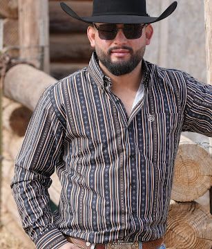 Man in black cowboy hat and sunglasses wearing a striped shirt, standing by stacked logs.