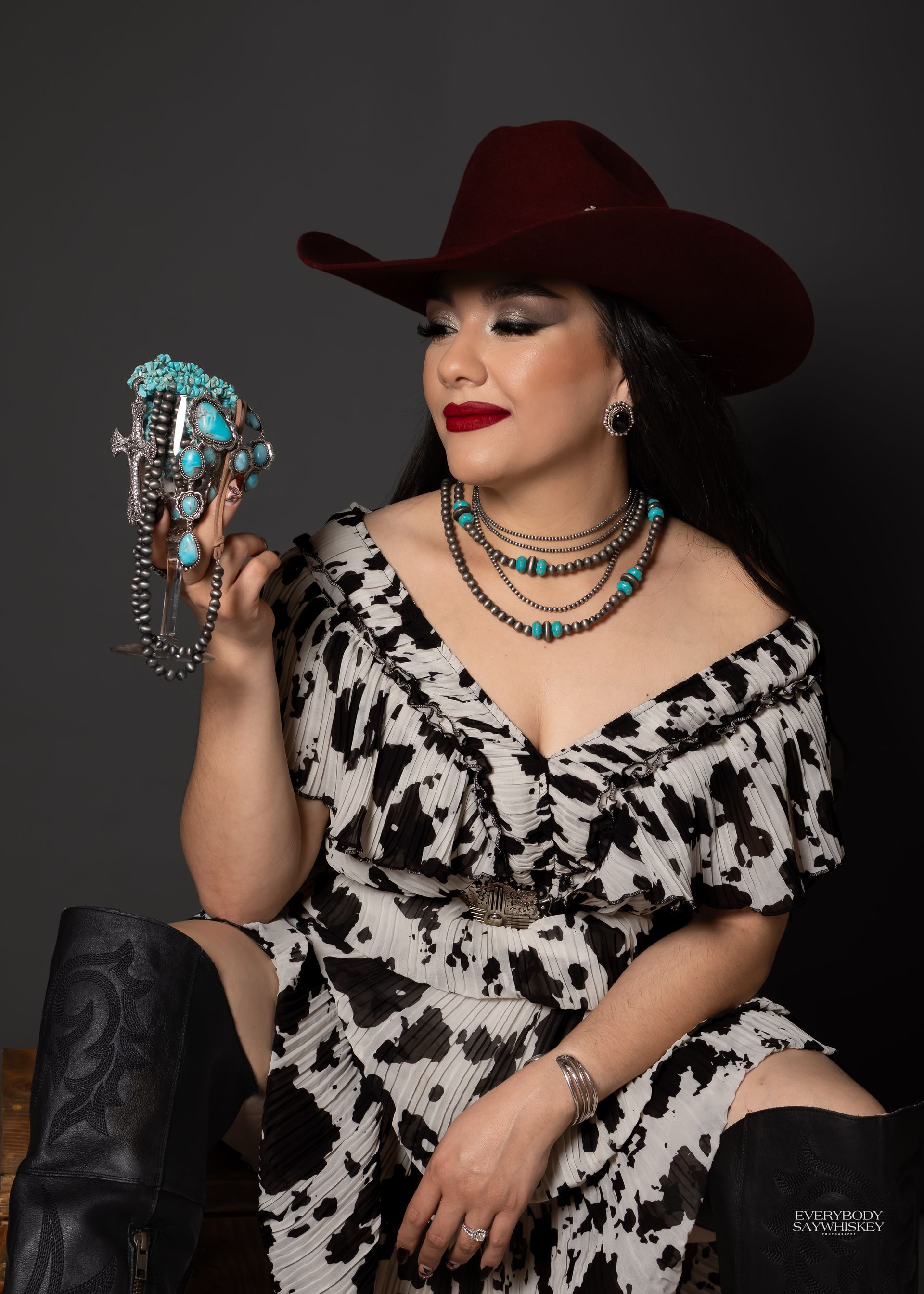 Woman in a black-and-white dress and red cowboy hat, holding a jeweled skull against a dark backdrop.