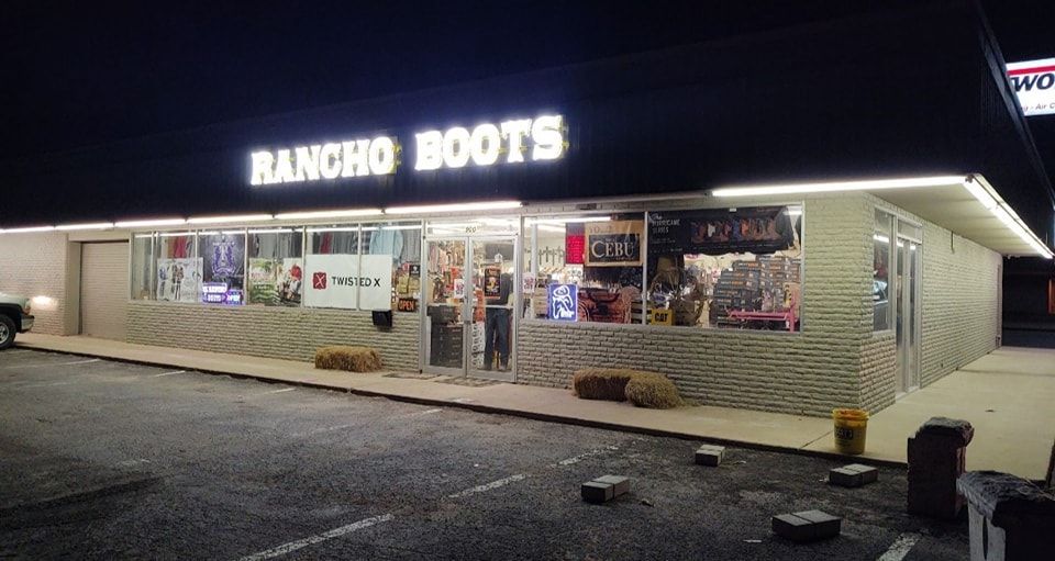 Rancho Boots storefront at night, lit sign above a gravel parking lot.