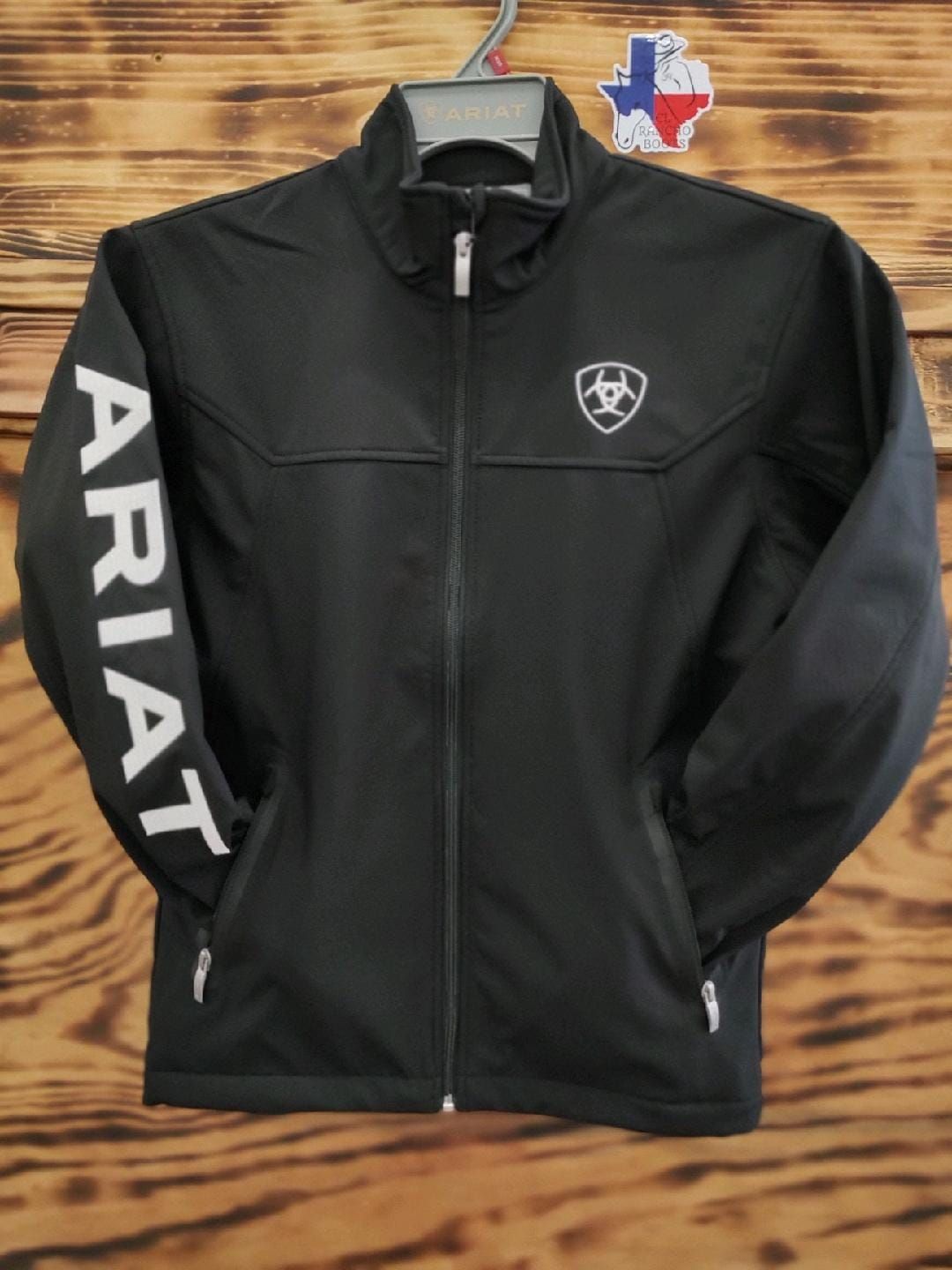 Black Ariat zip-up jacket with white sleeve logo, hanging on a hanger against a wood wall.