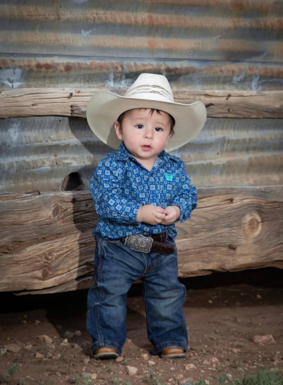 Child in a white cowboy hat, blue shirt, jeans, and boots standing by a weathered wooden wall.