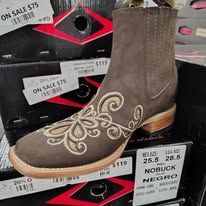 Brown nubuck boot with white swirl embroidery displayed on a store shelf with a price tag.