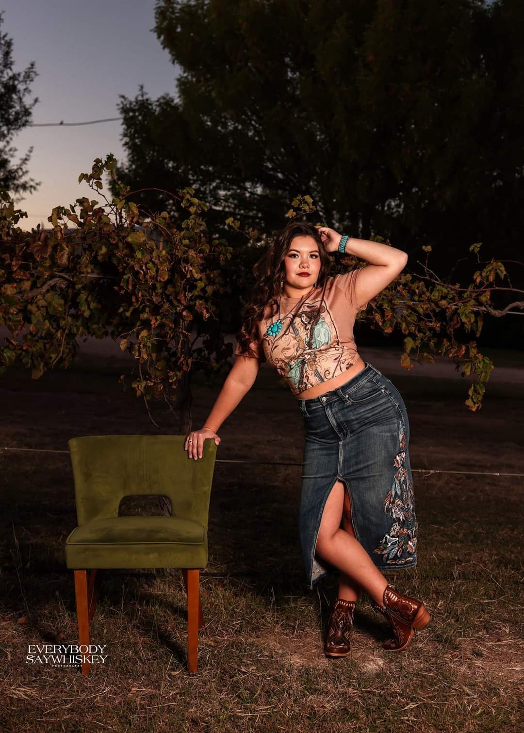 Woman posing outdoors beside a green chair at dusk, wearing a patterned top and denim skirt.
