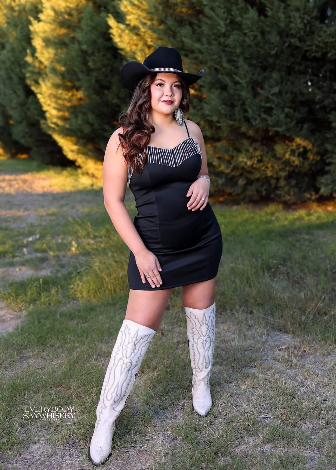Woman in a black dress and white boots posing outdoors in a grassy area, wearing a black hat.