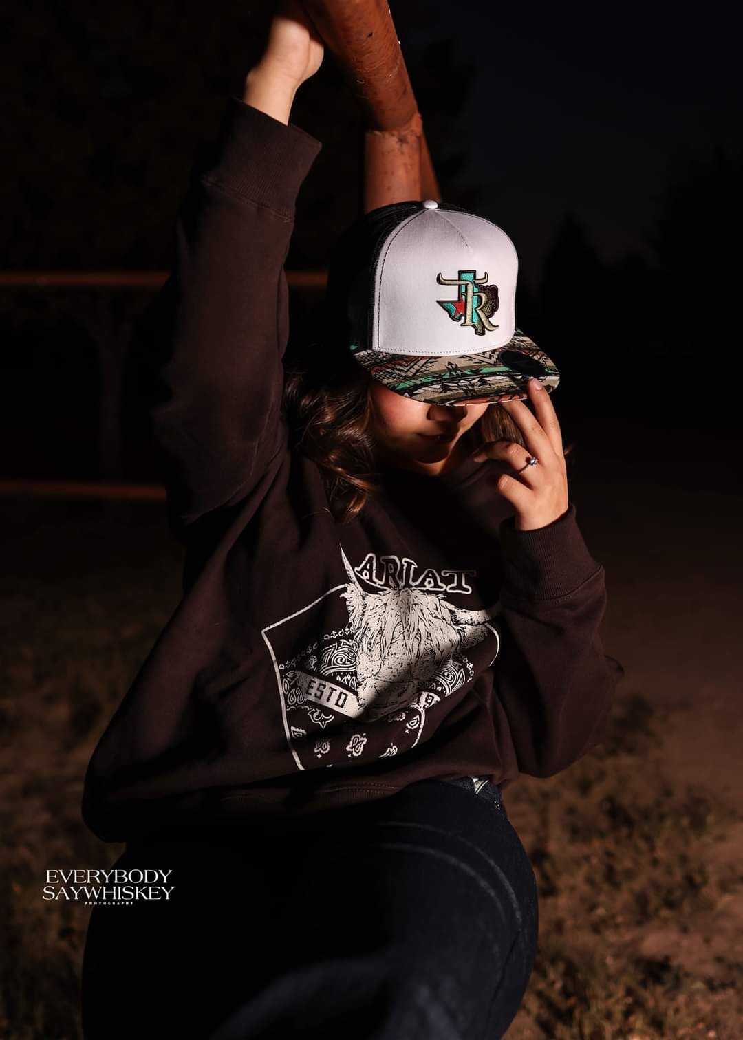 Person in a white cap and dark hoodie posing outdoors at night with one arm raised.