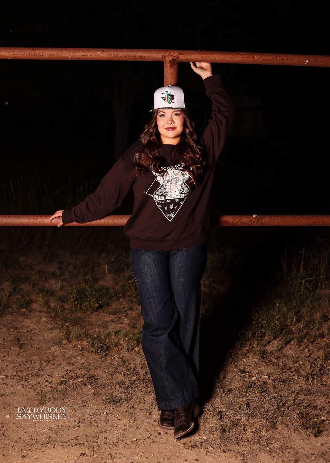 Person leaning against a wooden fence at night, wearing a dark hoodie and white cap.
