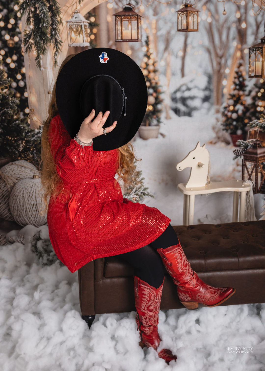 Woman in a red dress and cowboy boots sitting in a snowy holiday scene with lanterns and a black hat