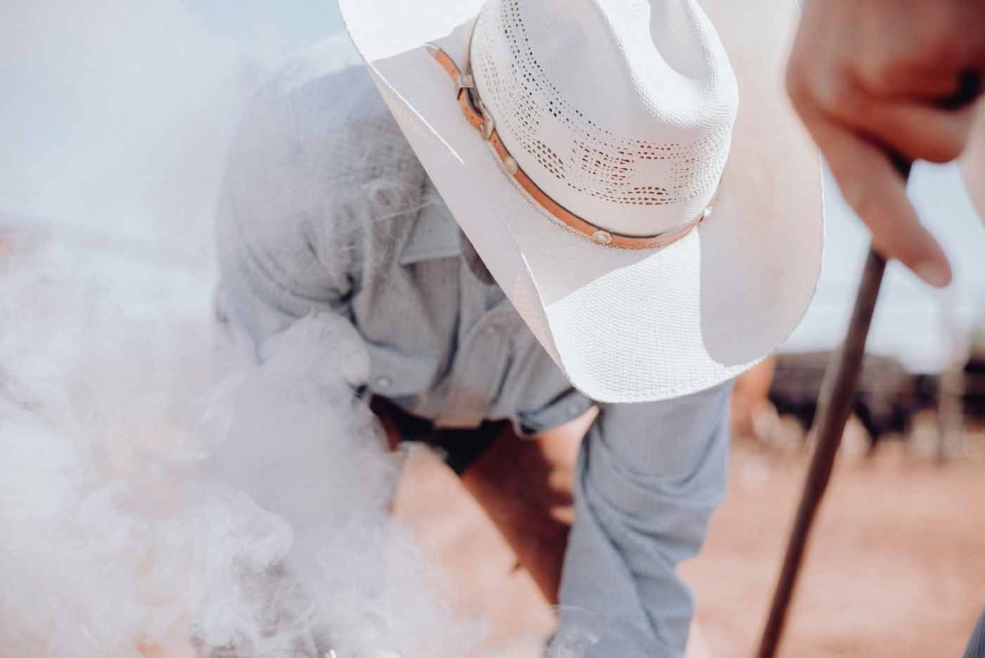 Cowboy in white hat bending over near a smoky livestock pen, holding a rope.