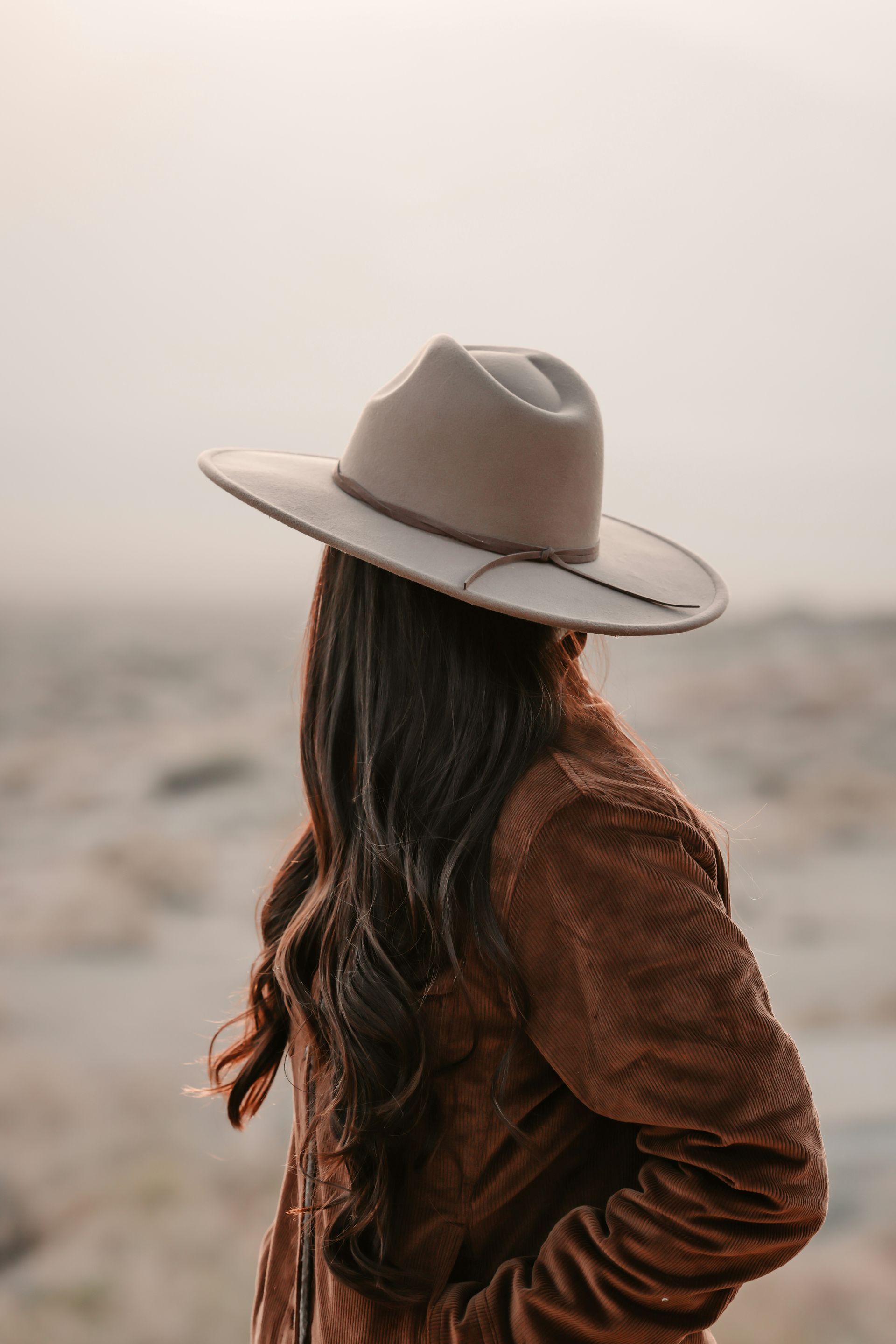 Person in a tan hat and brown jacket standing in a misty field, viewed from behind.