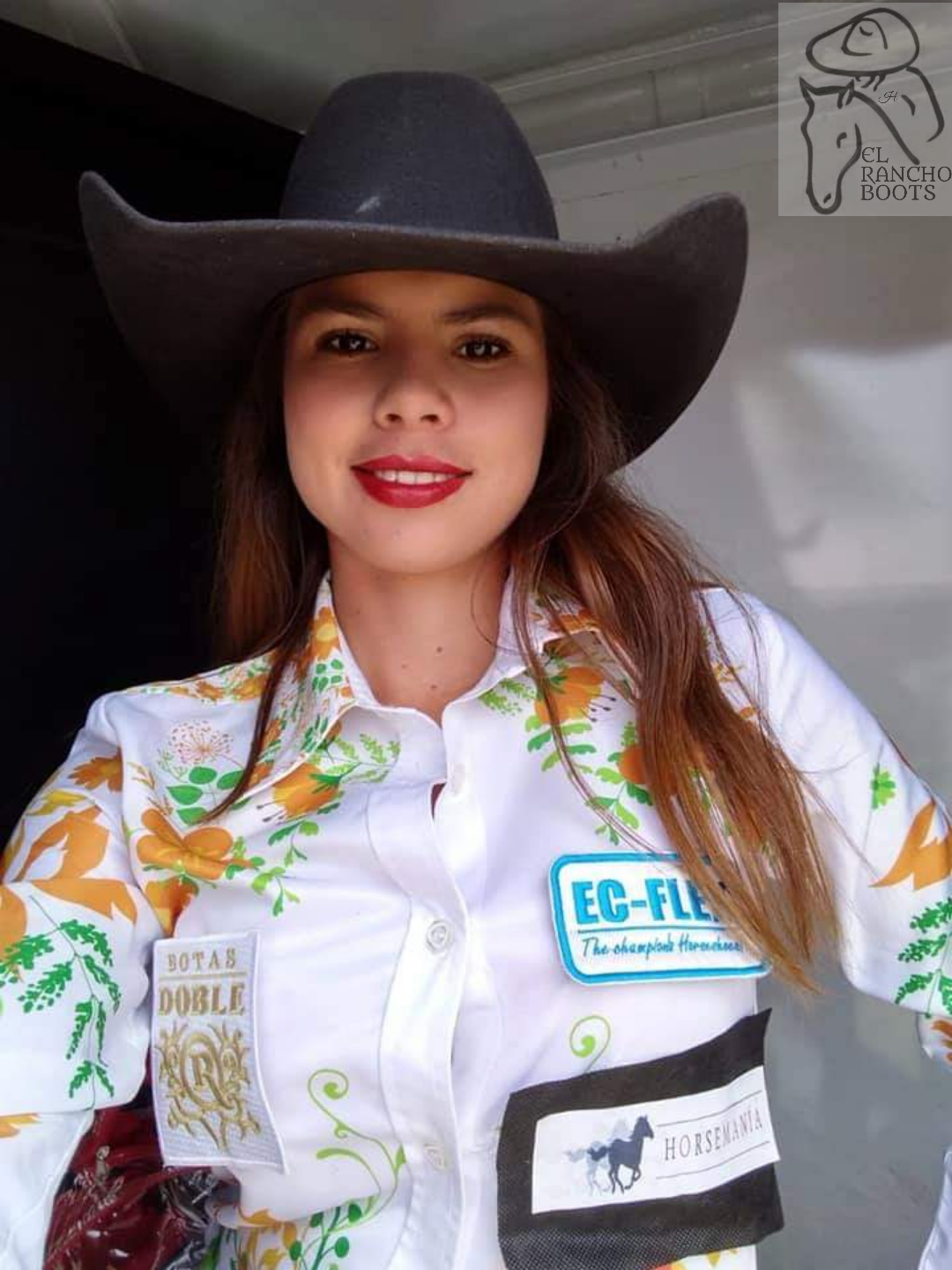 Woman in a black cowboy hat and white embroidered shirt, smiling indoors at the camera.