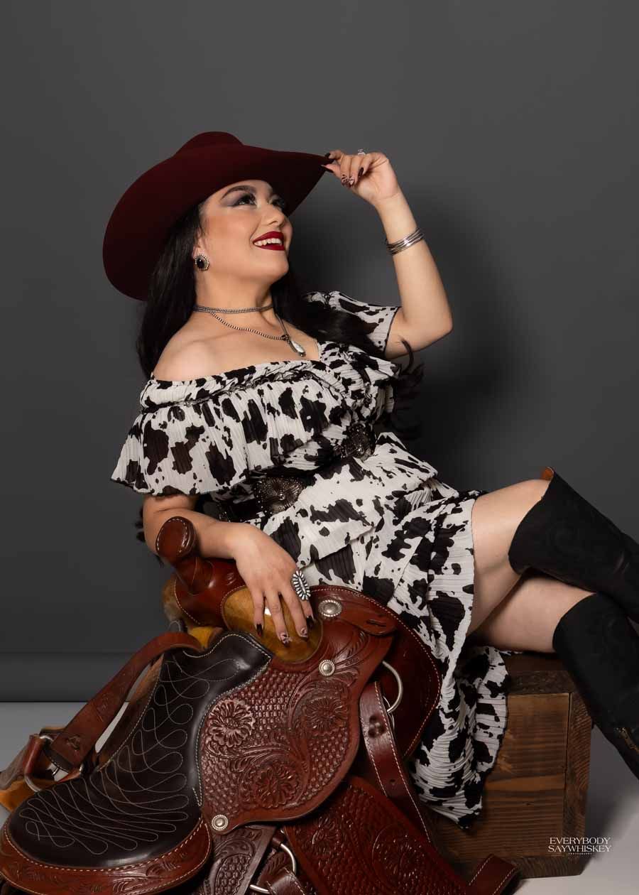 Woman in a burgundy hat posing on a carved leather saddle, wearing an off-shoulder black-and-white dress and black boots