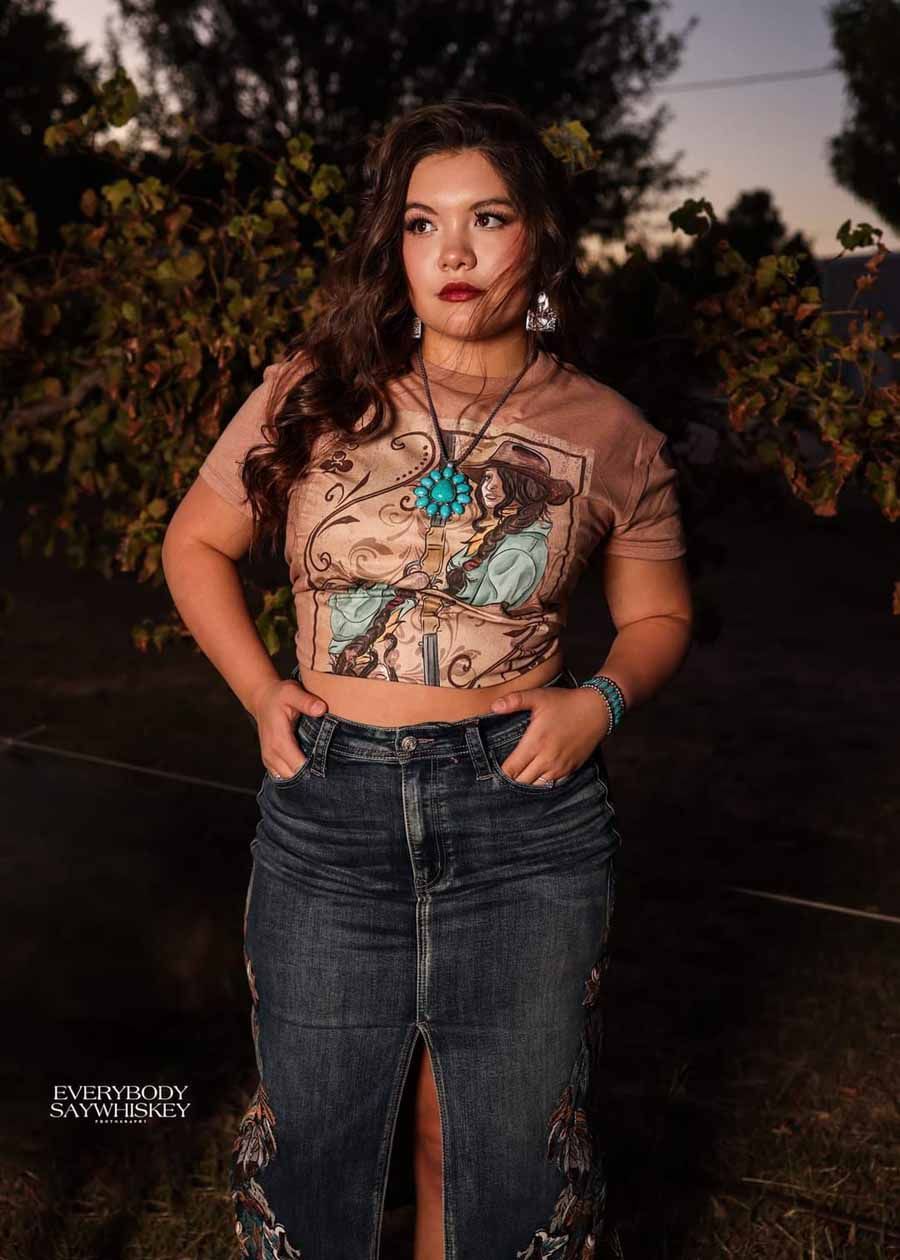 Person posing outdoors at dusk in a turquoise crop top and jeans, hand on hip.