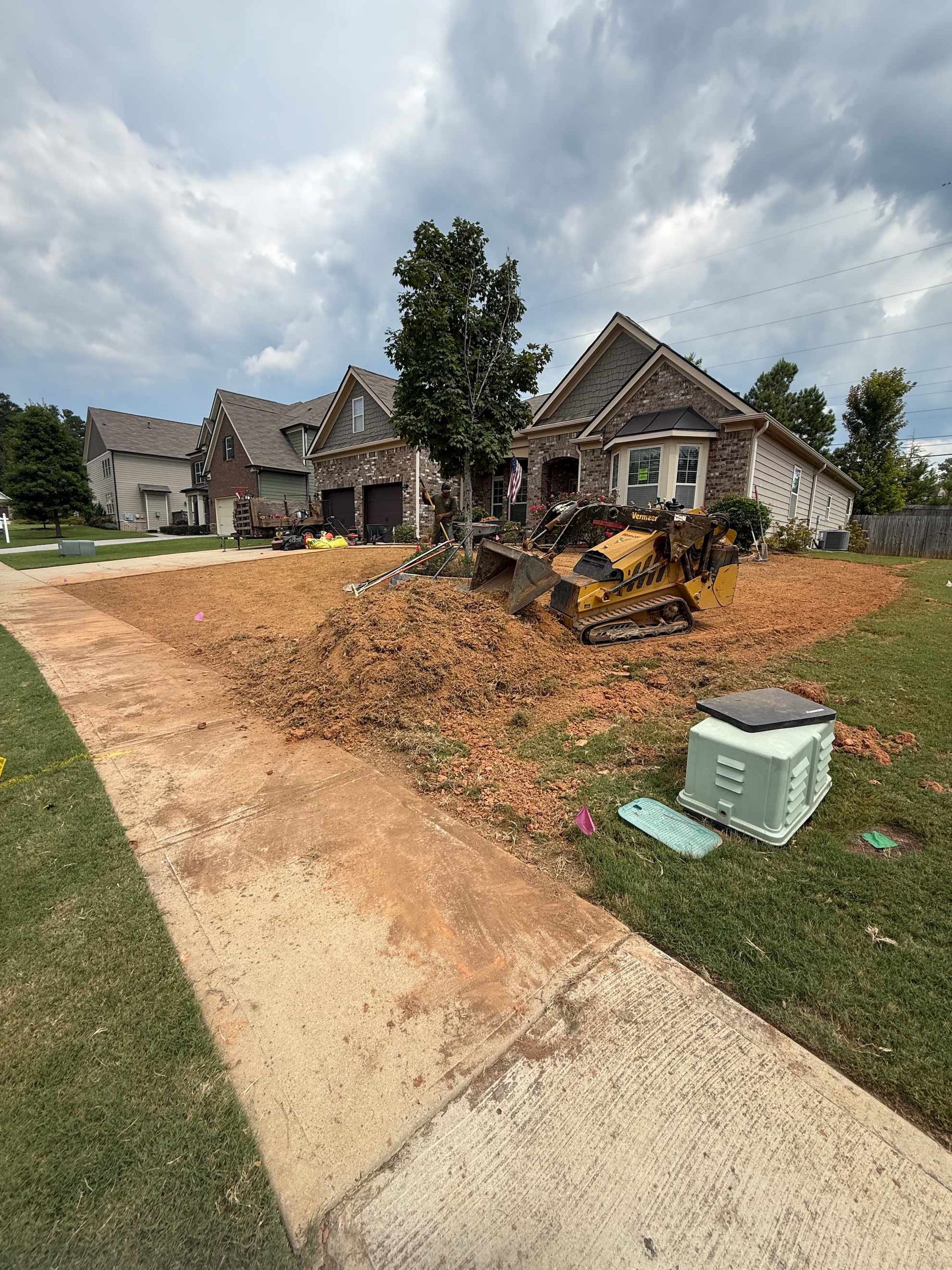 Construction in front yard of brick house, small bulldozer on dirt pile. Cloudy sky.