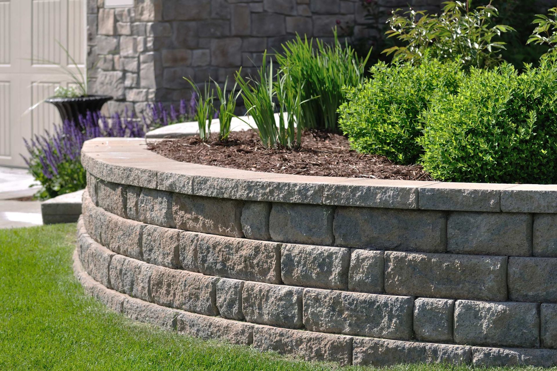 Curved retaining wall made of gray blocks, holding a flower bed with greenery. Green grass surrounds.