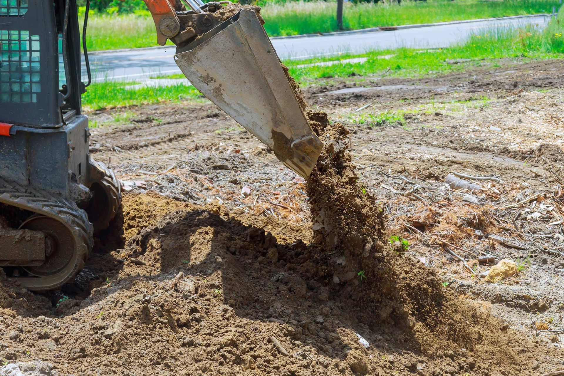 A small, tracked excavator dumping dirt into a ditch outdoors.