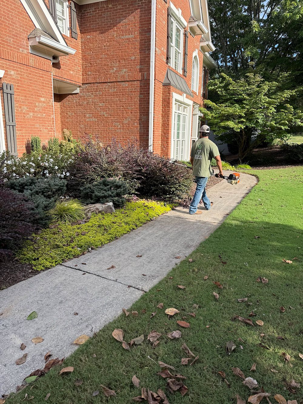 Man trimming sidewalk edges near a brick building with landscaped flowerbeds and green grass.