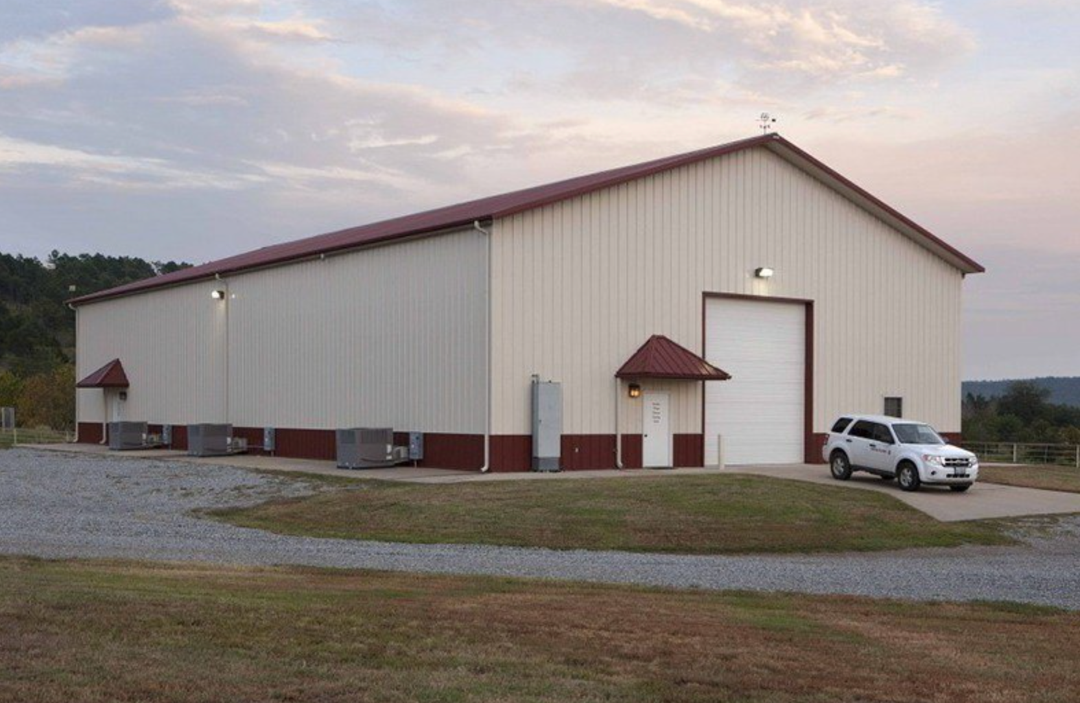Large beige metal building with a red roof and a parked white SUV on a gravel lot at dusk