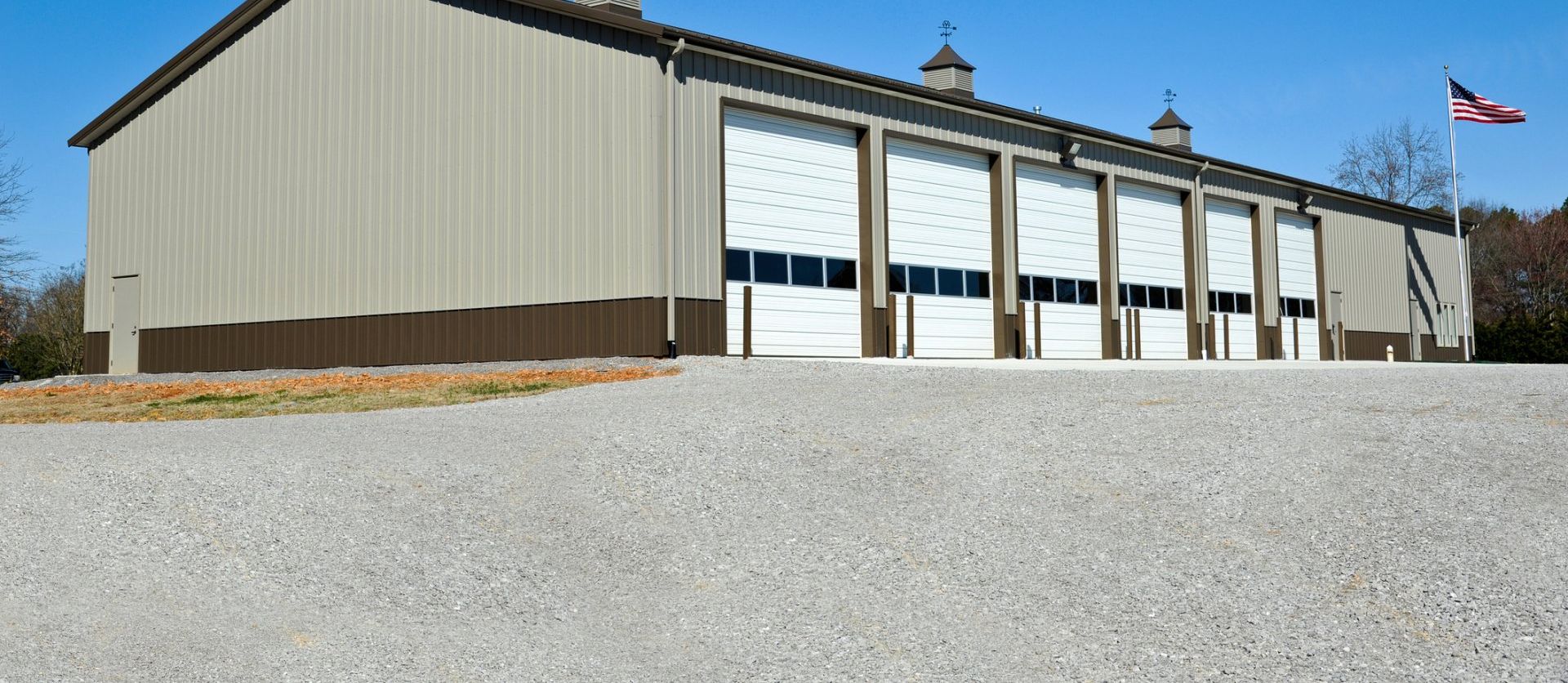 Large beige fire station with white garage doors and a flag flying on a gravel lot