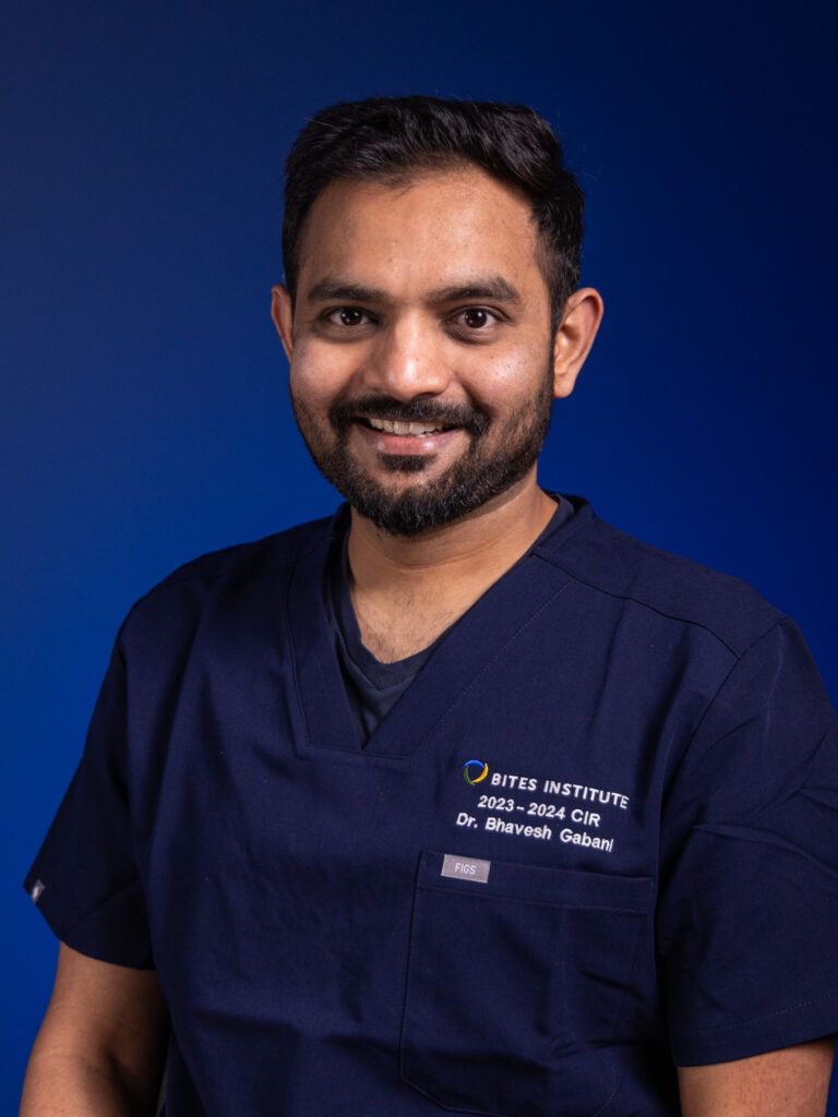 Man with dark hair and beard, wearing blue scrubs, smiling. Blue background.