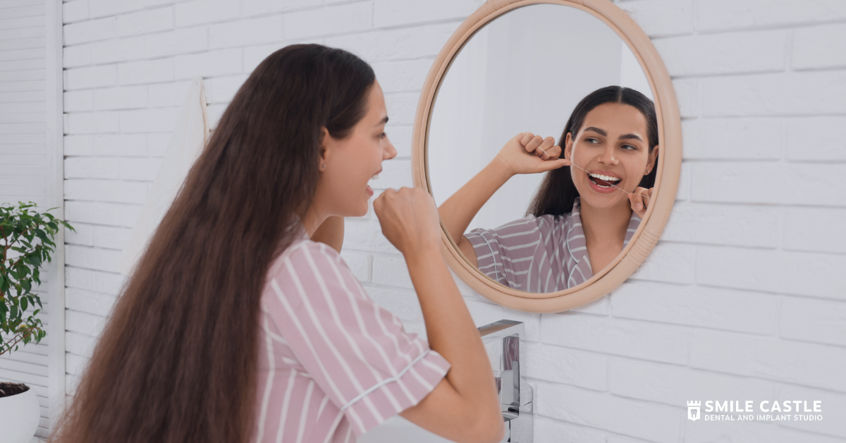 Woman flossing teeth while looking in a mirror in a bright bathroom.