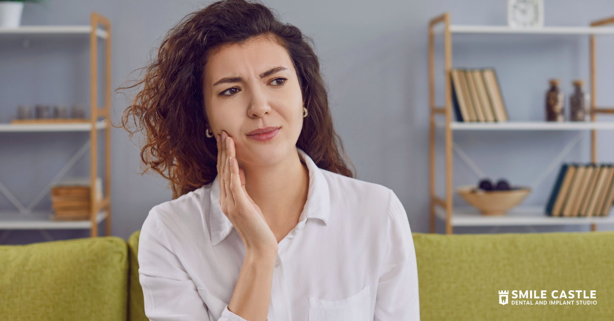 Woman in white shirt touching her cheek, sitting on a couch in a bright living room.