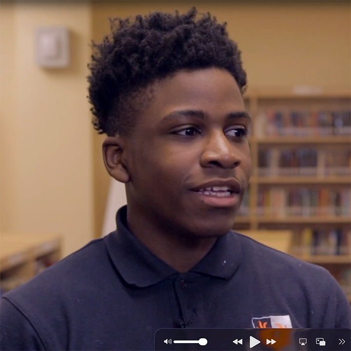 Young person with short, dark hair, wearing a dark polo shirt, speaking in a library setting.