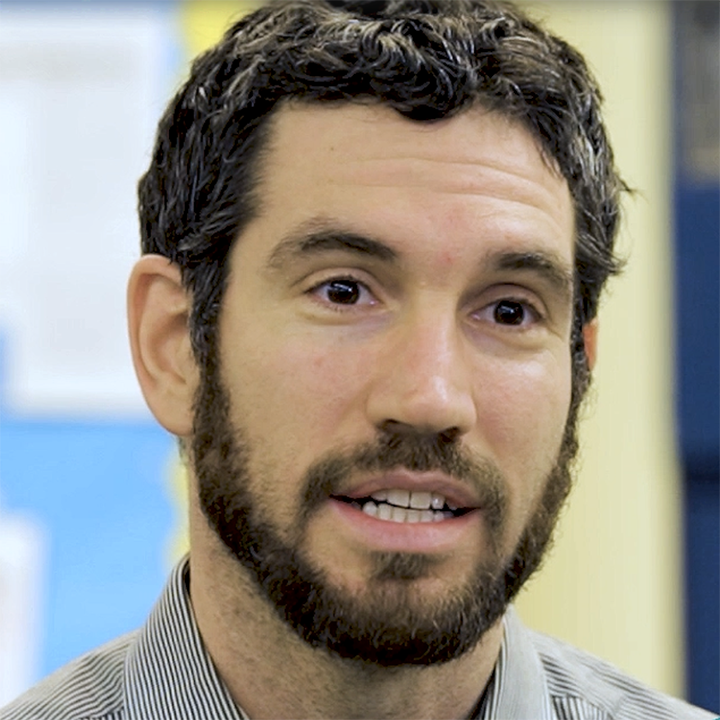 Man with brown curly hair, beard, and striped shirt, speaking indoors.