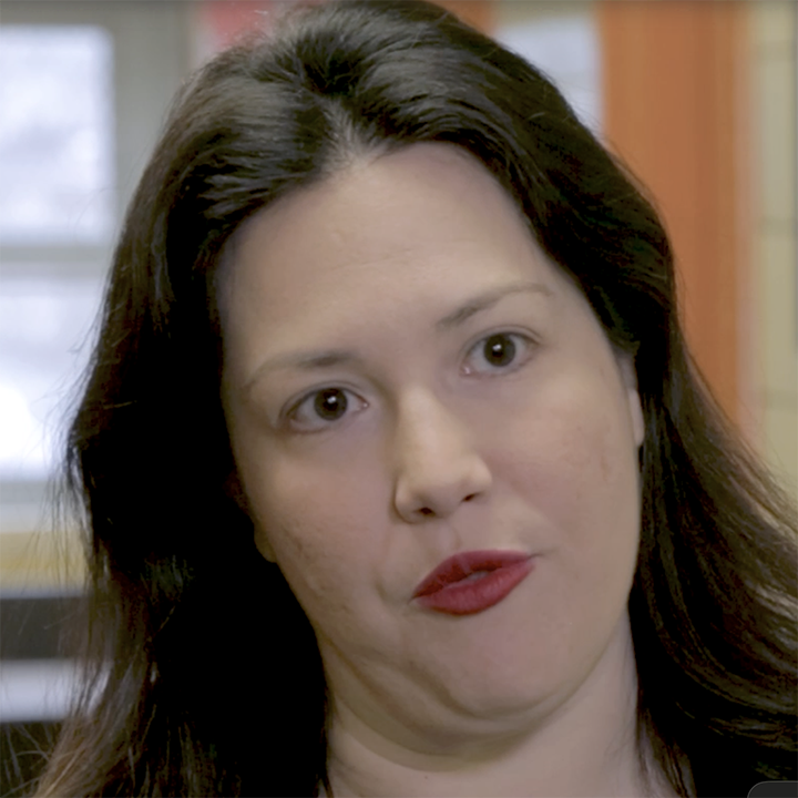 Woman with dark hair and red lipstick, looking towards the camera, indoor setting.
