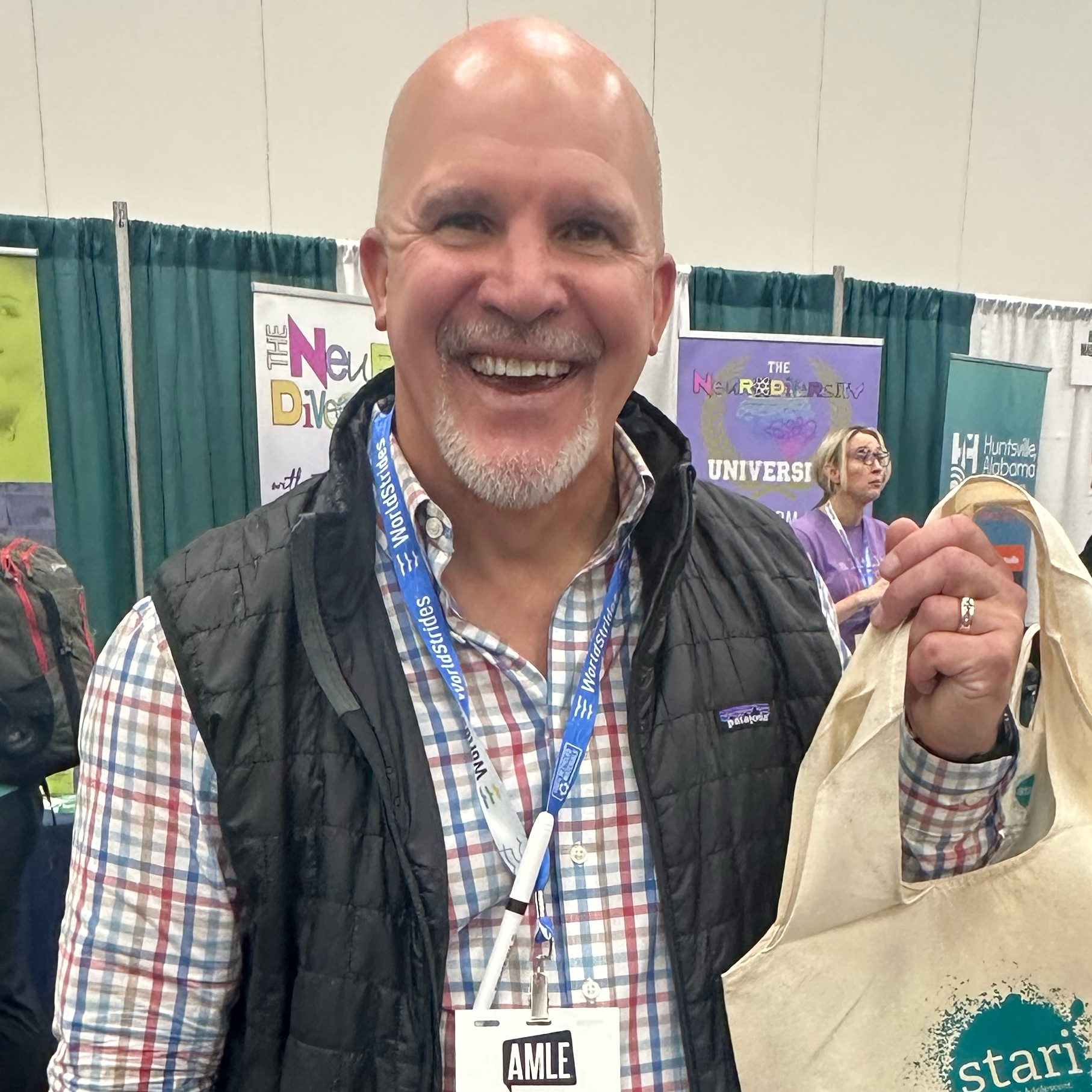 Man smiling, holding a tote bag, wearing a plaid shirt and vest, at a conference booth.