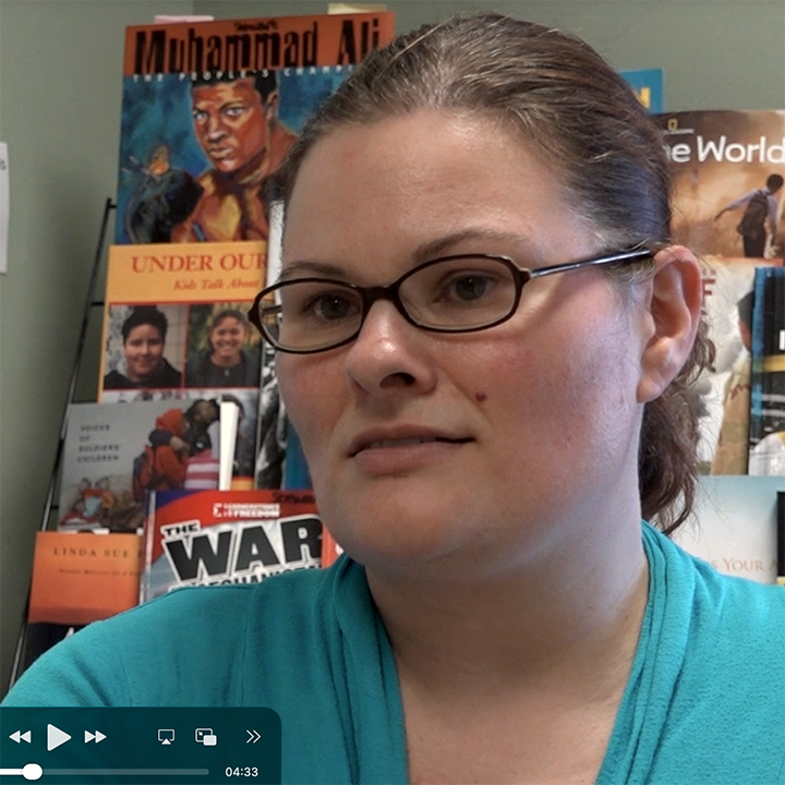 Woman with glasses in front of a bookshelf, wearing a teal top, looking slightly off-camera.