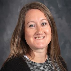 Woman with light brown hair smiles, wearing black cardigan over a gray and white patterned shirt.