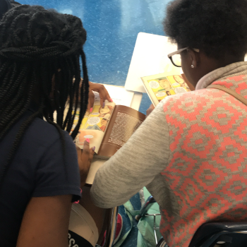 Two people, reading a book together indoors. One wears glasses and a patterned sweater.
