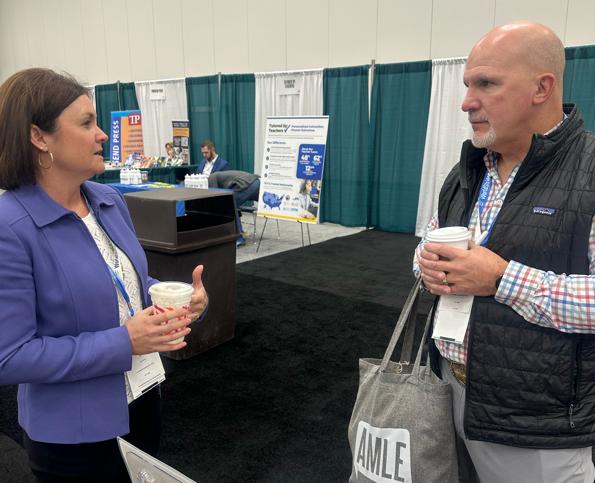 Two people converse at a conference booth, each holding a cup. Man wears a vest and plaid shirt. Woman gestures.