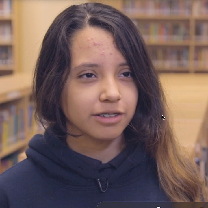 Young person with long hair wearing a dark blue hoodie, speaking in a library.