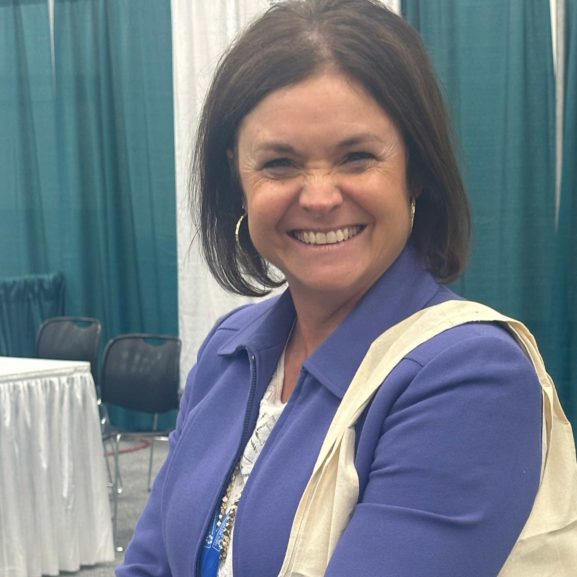 Woman smiles, wearing blue jacket and carrying a tote bag, indoors.