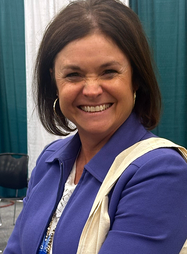 Woman smiling, wearing a blue jacket and hoop earrings, carrying a cream tote bag.