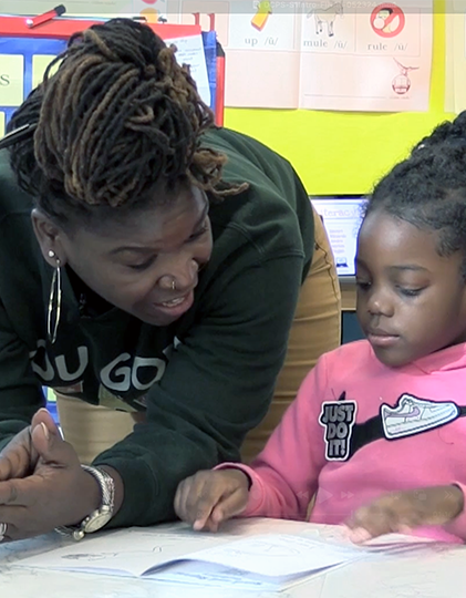 Teacher helping a student with reading. The teacher leans in, pointing at the page. The girl looks down at the paper.