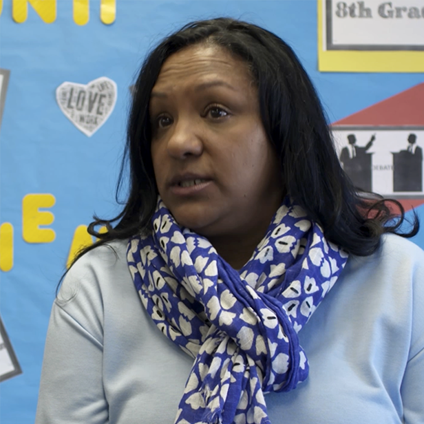 Woman in light blue shirt and blue patterned scarf speaking in front of a bulletin board.