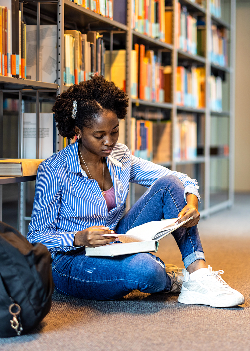 Woman reading a book on library floor in front of bookshelves, wearing blue jeans and a patterned shirt.