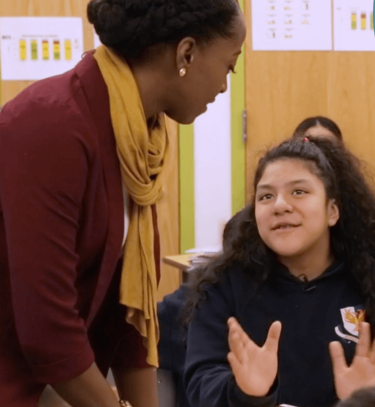 Teacher leans toward student in a classroom, student gestures.