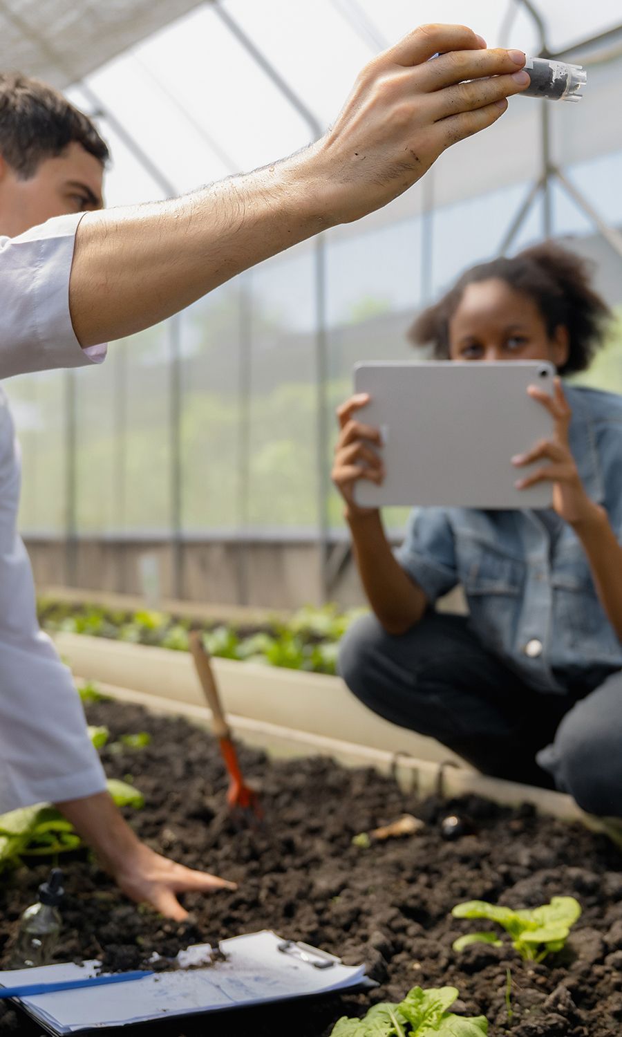 Person holding device over soil as another kneels, holding tablet in greenhouse.