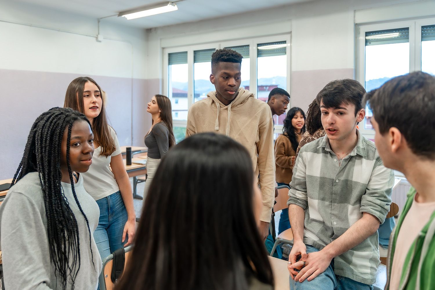 Students in a classroom, talking in small groups. Some are standing, and some are sitting near desks.