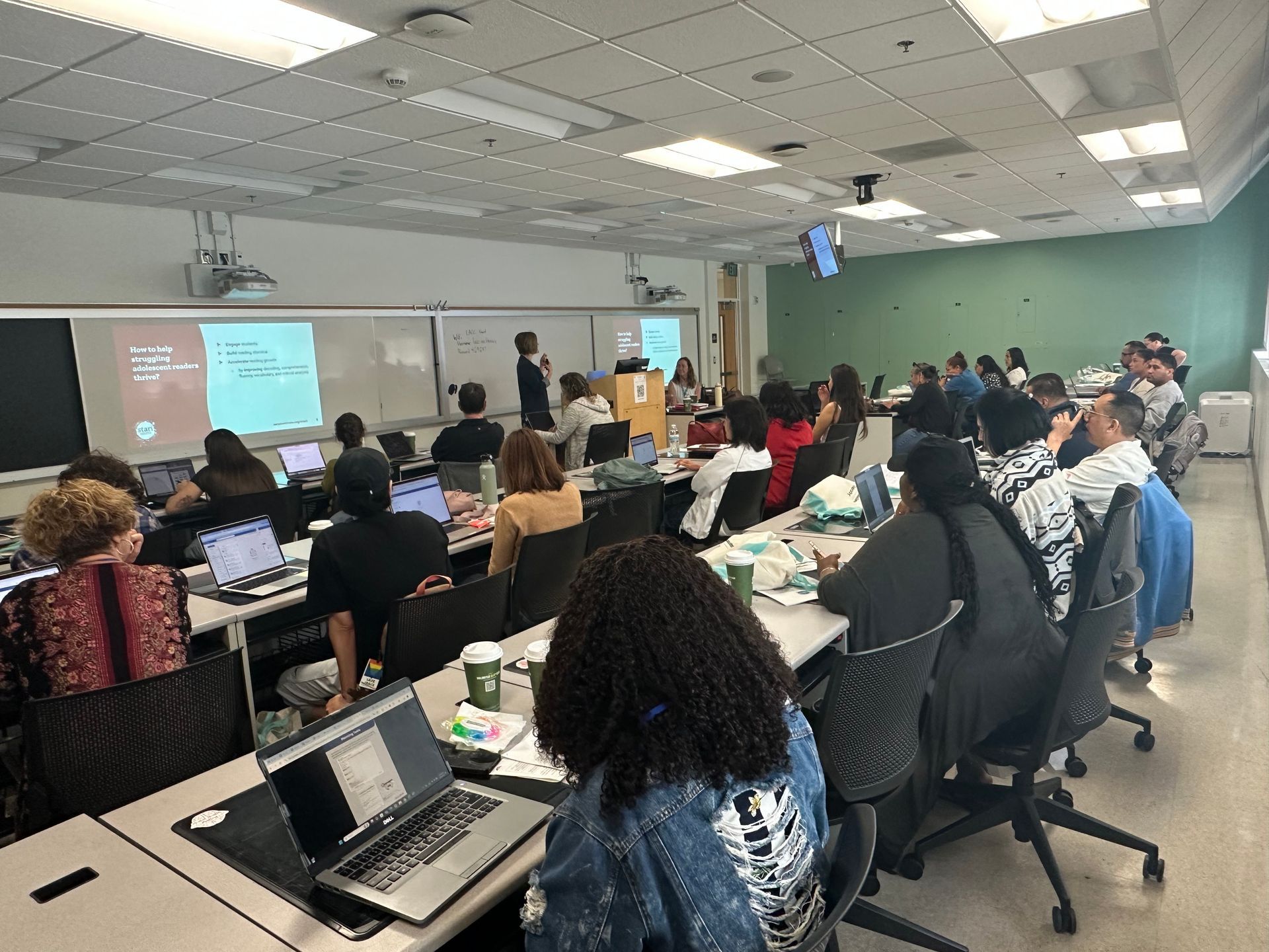 Students in a classroom, some working on laptops. A presenter stands at the front by a screen with text.