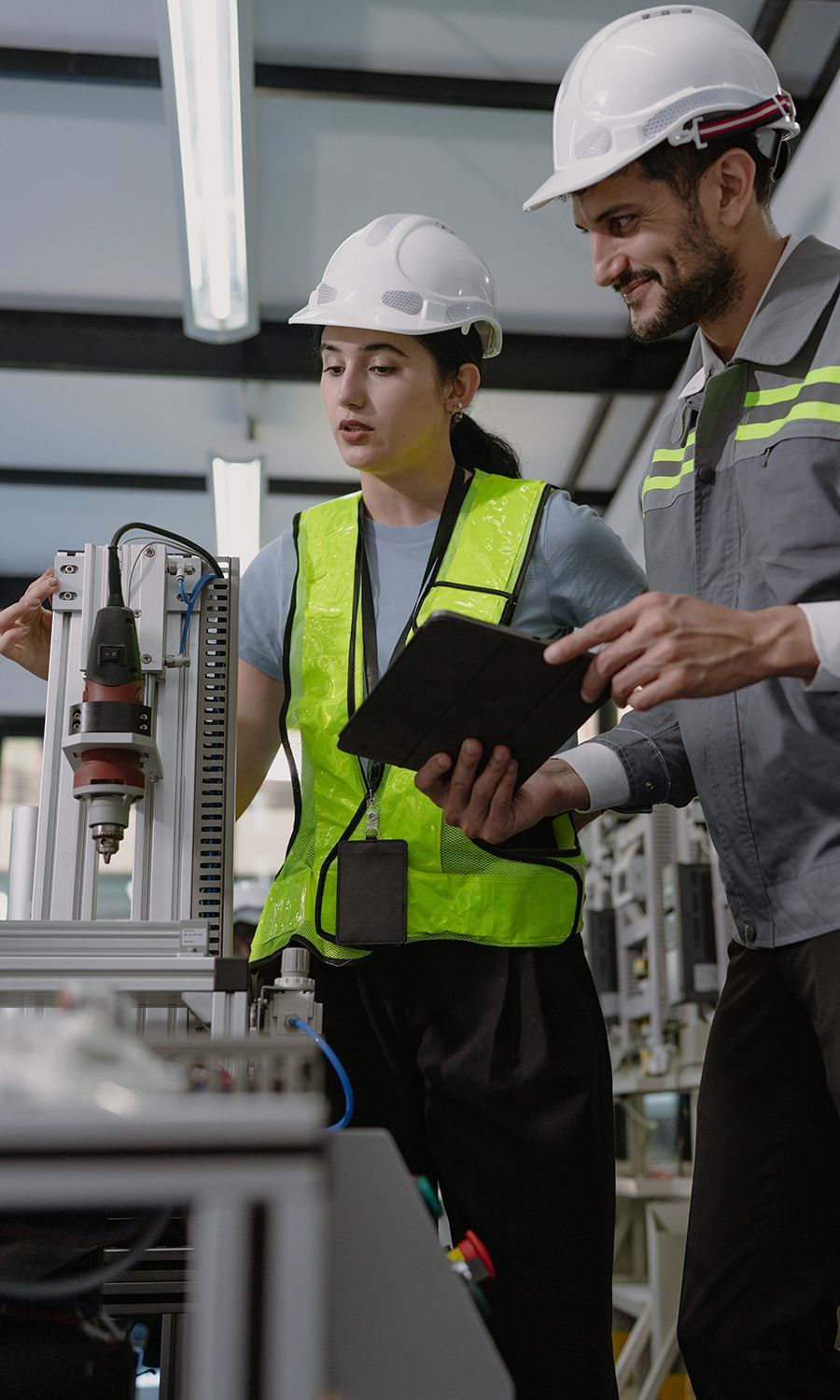 Two people wearing hard hats and safety vests examining machinery in a factory.
