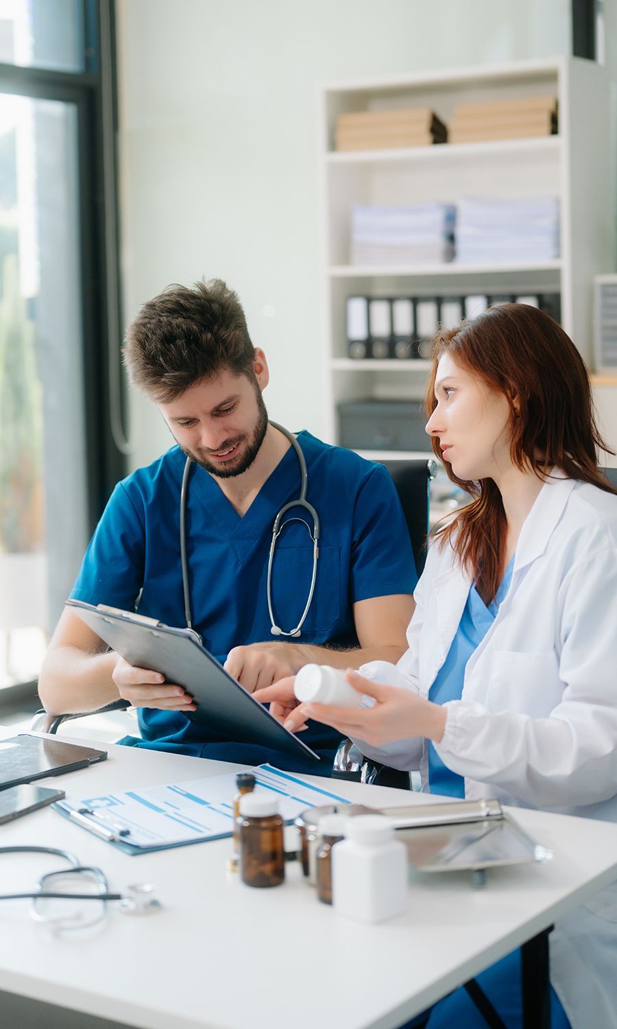 Two healthcare workers reviewing documents at a desk; man in scrubs, woman in lab coat holding medication.