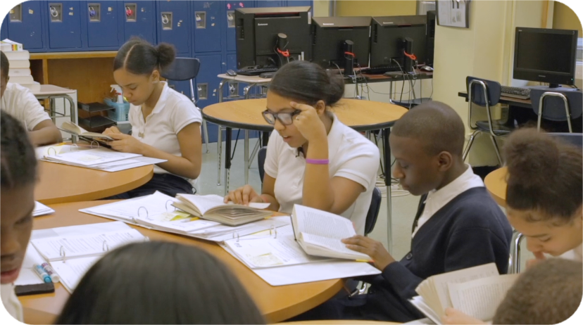 Students in a classroom, seated around tables, reading books and writing. Computers in the background.